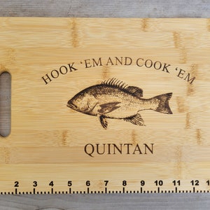 May include: A bamboo chopping board with a fish design and the text "HOOK 'EM AND COOK 'EM" and "QUINTAN". The board has a handle and a ruler on the bottom edge.