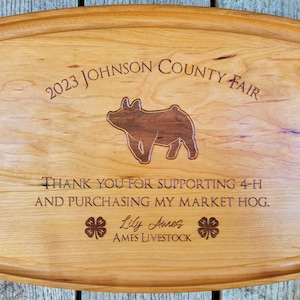 May include: Wooden serving tray with a carved pig silhouette and text that reads "2023 Johnson County Fair." The tray also includes the text "Thank you for supporting 4-H and purchasing my market hog." and the name "Lily Aures."