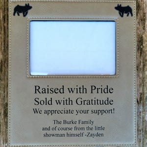 Puede incluir: Un marco de fotos rectangular de piel sintética beige con costuras blancas. Las esquinas superiores presentan siluetas de cerdos negros. El texto central dice: Raised with Pride, Sold with Gratitude, We appreciate your support! The Burke Family and of course from the little showman himself -Zayden.