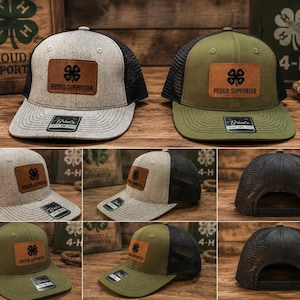 Puede incluir: Dos gorras tipo camionero con un parche de cuero marrón que muestra un trébol de cuatro hojas negro y el texto PROUD SUPPORTER. Una gorra es gris jaspeado con malla negra y la otra es verde oliva con malla negra. Ambas tienen visera plana.