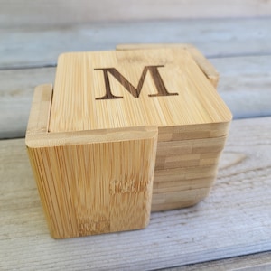 Puede incluir: Un juego de posavasos cuadrados de bambú con un soporte. Cada posavasos está grabado con la letra "M". Los posavasos y el soporte están hechos de bambú de color claro, con la veta de la madera visible.