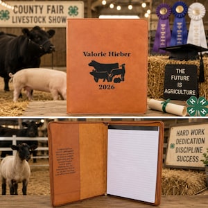 May include: A tan leather portfolio featuring the name Valorie Hieber, a graphic of a cow, pig, and sheep, and the year 2026. The interior includes a lined notepad and a printed message on the left side, with a smooth, durable exterior finish.