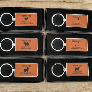 May include: Six black boxes with orange rectangular keychains inside. Each keychain has a different animal silhouette and the text "Thank You for Buying My 2025 Market [Animal]"