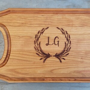 Puede incluir: Una tabla de cortar de madera con un asa y un diseño decorativo de una corona con las iniciales "LG" en el centro.