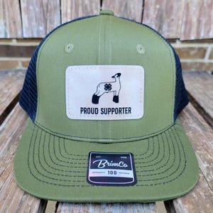 May include: Olive green and navy blue trucker hat with a patch featuring a sheep and a four-leaf clover, with the words "PROUD SUPPORTER". The hat also has a BrimCo label.