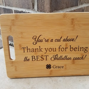 Könnte beinhalten: Ein braunes Bambus-Schneidebrett mit einem Griff. Das Brett ist mit dem Text "You're a cut above! Thank you for being the BEST Thillathon coach! Grace" und einem vierblättrigen Kleeblatt graviert.