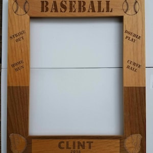 May include: A wooden picture frame with a baseball theme. The frame is made of light and dark wood and has a cutout of a baseball glove in each corner. The frame has the words "BASEBALL" at the top and "CLINT 2018" at the bottom. The frame is designed to hold a photo.