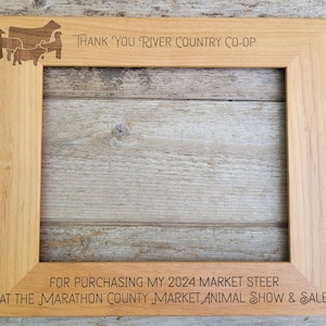 May include: A wooden picture frame with an engraved message thanking someone for purchasing a 2024 market steer at the Marathon County Market Animal Show & Sale. The frame has a silhouette of cattle in the top left corner.