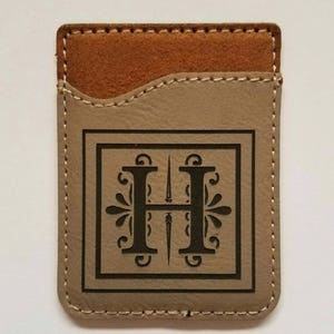 Puede incluir: Una cartera de teléfono de cuero marrón con un borde cosido y un bolsillo para una tarjeta de crédito. La cartera tiene un diseño grabado decorativo de la letra "H" en un marco cuadrado.