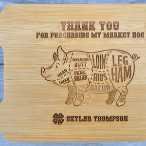 Puede incluir: Una tabla de cortar de madera con una ilustración de cerdo que muestra diferentes cortes de carne. El texto "THANK YOU FOR PURCHASING MY MARKET HOG" está grabado en la tabla. La tabla también tiene el texto "SKYLAR THOMPSON" y un trébol de cuatro hojas.