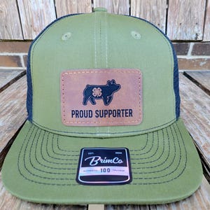 Puede incluir: Gorra de camionero verde oliva con parte trasera de malla azul marino y visera curva. Un parche de cuero marrón presenta un cerdo negro con un trébol de cuatro hojas y las palabras "PROUD SUPPORTER". La gorra también tiene una etiqueta BrimCo.