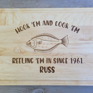 May include: A wooden cutting board with an engraved image of a fish and the text "Hook 'em and cook 'em. Reeling 'em in since 1961. Russ."