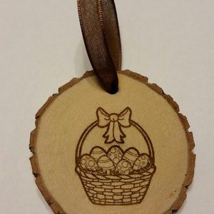 May include: A wooden Easter ornament with a brown ribbon. The ornament is a round slice of wood with a laser-engraved design of a basket filled with Easter eggs and a bow on top.