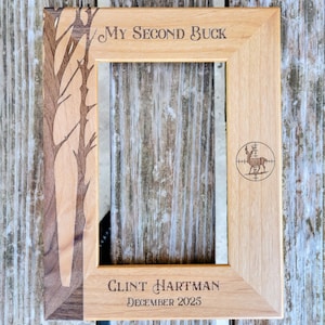 Könnte beinhalten: Heller Holzbilderrahmen mit dem Text "My Second Buck" und einer Hirschzielscheibe. Der Rahmen hat ein Baumdesign auf der linken Seite und den Namen "Clint Hartman" sowie das Datum "Dezember 2025" unten.