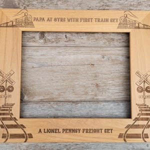 Op de afbeelding: Een houten fotolijst met een treindesigne. De lijst heeft een uitsparing voor een foto en is gegraveerd met de tekst "Papa at 8 yrs with first train set" en "A Lionel Pennsy Freight Set".
