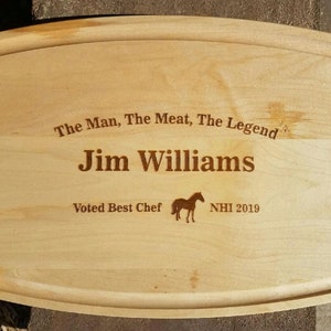Op de afbeelding: Een lichtgekleurde houten snijplank met een gebogen rechthoekige vorm. De tekst "The Man, The Meat, The Legend" en "Jim Williams" zijn gegraveerd. Daaronder staat "Voted Best Chef" en een paardenafbeelding met "NHI 2019".