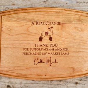 Puede incluir: Una tabla de cortar de madera con un mensaje grabado que dice "A Real Change Thank You for supporting 4-H and for purchasing my market lamb Callie Mecks". La tabla también presenta una imagen grabada de un cordero.