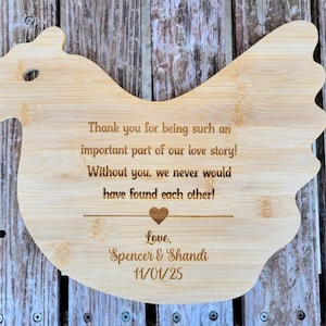 May include: A light-colored, chicken-shaped wooden cutting board with engraved text. The text reads, "Thank you for being such an important part of our love story! Without you, we never would have found each other!" with a heart and names.