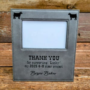 Puede incluir: Marco de fotos gris con apertura rectangular. El marco tiene siluetas de vacas negras en las esquinas y el texto "THANK YOU for supporting 'Lucky' my 2025 4-H steer project Bryar Baker."
