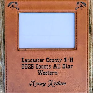 May include: A brown leather picture frame with a rectangular opening. The frame has a horse head etched in each upper corner. The text "Lancaster County 4-H 2025 County All Star Western Avery Kellum" is printed on the frame.