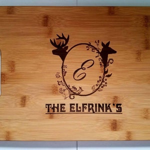 May include: A personalized bamboo cutting board with a monogrammed "E" surrounded by a floral design and two deer silhouettes. The text "THE ELFRINK'S" is engraved below the design.