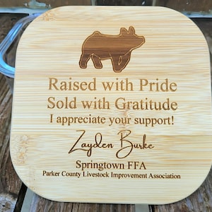 Può includere: Un coperchio quadrato in bambù con angoli arrotondati che presenta una silhouette di maiale incisa al laser sulla parte superiore. Il testo recita: Raised with Pride, Sold with Gratitude, I appreciate your support!, Zayden Burke, Springtown FFA, Parker County Livestock Improvement Association.