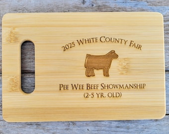 May include: A wooden cutting board with a handle, engraved with the text "2025 White County Fair Pee Wee Beef Showmanship (2-5 yr. old)" and a silhouette of a cow.