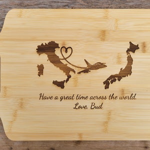 May include: A wooden cutting board with a laser-engraved design of Italy and Japan connected by a plane and a heart. The text "Have a great time across the world. Love, Bud" is engraved below the design.