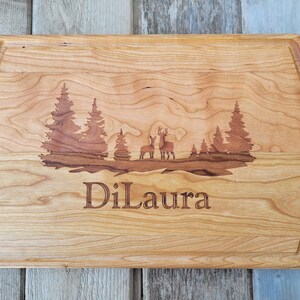 May include: A light-colored wooden cutting board with a carved forest scene featuring deer and trees. The name "DiLaura" is engraved below the scene. The board has a juice groove around the edge.