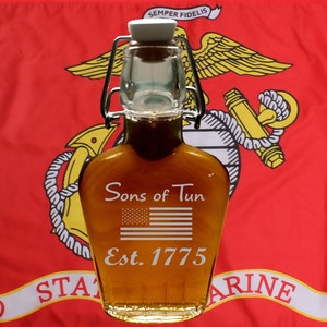 May include: A glass bottle with a metal swing top lid filled with a dark amber liquid. The bottle is decorated with the text "Sons of Gun" and a small American flag. The text "Est. 1775" is also engraved on the bottle. The bottle is in front of a red, white, and yellow United States Marine Corps flag.