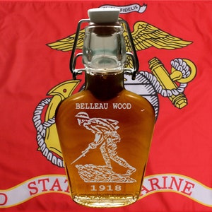 May include: A glass bottle with a metal swing top lid filled with a golden liquid. The bottle has an etched image of a soldier in a military uniform with the text "Belleau Wood 1918". The bottle is in front of a red flag with a yellow emblem.