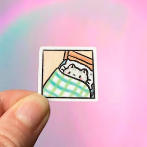 May include: A small, square sticker featuring a cartoon cat tucked into bed. The cat is under a green and white checkered blanket. The sticker has a white border and is held against a pink and rainbow background.