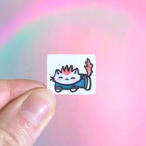 May include: A small, square sticker featuring a cartoon cat with pink and red accents. The cat is wearing a blue outfit and has a spiky hairstyle. The sticker is held between fingers against a pink and iridescent background.