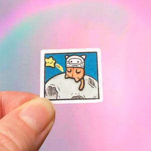 May include: A small, square sticker featuring a cartoon cat wearing a space helmet, resting on a moon. A yellow star with a trail is in the upper left corner. The background is blue, and the sticker is held by a hand.