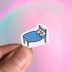 May include: A sticker featuring a cartoon bed with a blue blanket and an angry face. The bed has a wooden frame and legs. The background is a soft pink with a rainbow effect.