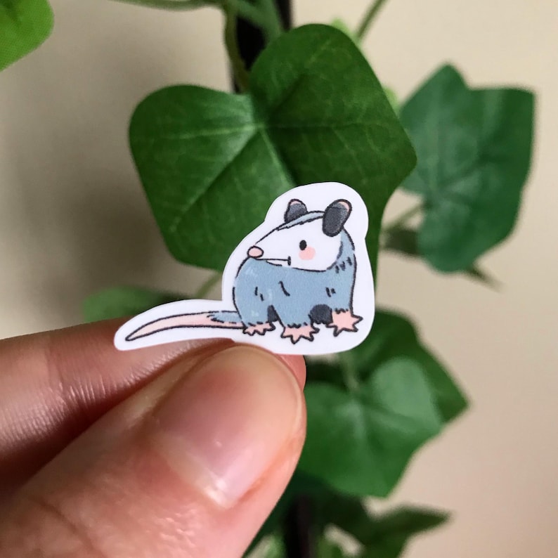 Pode incluir: Um adesivo com um desenho de um gamb&aacute;. O gamb&aacute; tem um corpo azul claro, bochechas rosadas e uma cauda longa e fina. O adesivo &eacute; segurado pelos dedos de uma pessoa, com folhas verdes ao fundo.