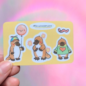 Puede incluir: Una hoja de pegatinas con dibujos animados de ornitorrincos con diferentes expresiones y accesorios. Las pegatinas están sobre un fondo amarillo con el texto "@AnnalisesArtwork" en la parte superior.