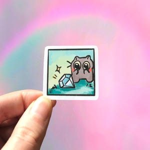 May include: A square sticker featuring a cartoon illustration of a gray creature with large eyes and a blushing face, looking at a sparkling blue diamond. The background is a light blue square with green accents. The sticker is held against a pink and rainbow background.