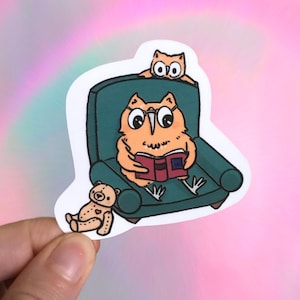 May include: A sticker featuring an orange owl wearing glasses, reading a red book while seated in a teal armchair. A smaller owl sits behind the chair, and a teddy bear is at the owl's feet. The background is a gradient of pink and blue.