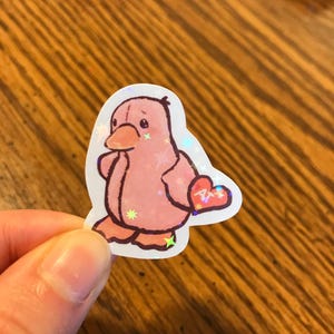 以下が含まれることがあります： ハート型の尾を持つ漫画のアヒルのピンク色のステッカー。ステッカーは星型のアクセントが付いたホログラフィック仕上げです。アヒルは翼を顔に当てています。ステッカーは人が持っています。