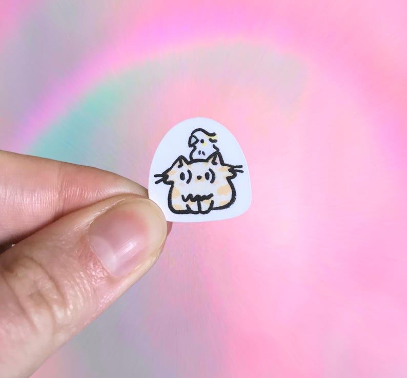Puede incluir: Una peque&ntilde;a pegatina blanca con una ilustraci&oacute;n de dibujos animados de un gato y un p&aacute;jaro. El gato tiene una cara redonda, bigotes y una expresi&oacute;n sonriente. El p&aacute;jaro tiene una peque&ntilde;a cresta. La pegatina est&aacute; sujeta por una mano.