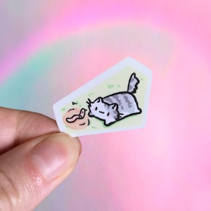 May include: A sticker featuring a cartoon cat with gray and white stripes, lying on green grass, looking at a worm. The cat has a happy expression. The sticker has a light yellow border and is set against a colorful, blurred background.