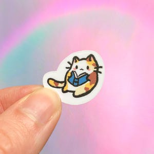 May include: A small, white sticker featuring a cartoon cat reading a blue book. The cat has orange, brown, and white fur with black outlines. The background is a soft pink and rainbow gradient.