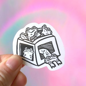 May include: A black and white sticker of a box with four frogs inside. The box is a cube shape with a lid. The frogs are all different sizes and have different expressions. The sticker is on a pink background.