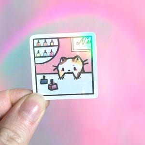 May include: A square holographic sticker featuring a cartoon calico cat behind a counter in a nail salon. The scene includes shelves with colorful nail polish bottles and two bottles on the counter. The sticker has a white border and black outlines.