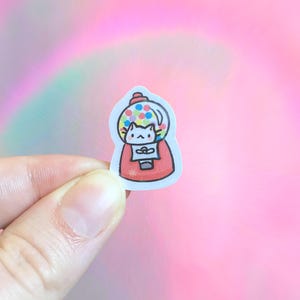 May include: A sticker featuring a cartoon gumball machine with a cat inside. The gumball machine is red with a clear top filled with colorful gumballs. The cat is white with black details. The background is a soft pink.