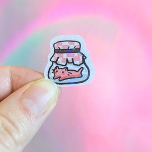 May include: A sticker featuring a cartoon cat inside a jar. The jar has a red and white checkered lid. The cat is pink and lying down. The sticker has a holographic effect, with a pink and blue background.