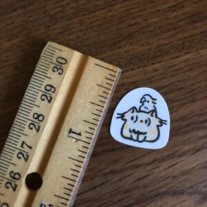 Puede incluir: Una peque&ntilde;a pegatina blanca con una ilustraci&oacute;n de dibujos animados de dos gatos y un p&aacute;jaro. Los gatos son de color naranja claro con contornos negros y el p&aacute;jaro es amarillo. Una regla de madera est&aacute; al lado de la pegatina.