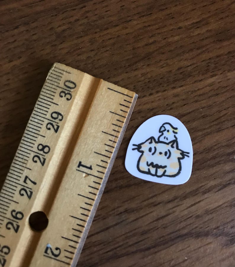 Puede incluir: Una peque&ntilde;a pegatina blanca con una ilustraci&oacute;n de dibujos animados de dos gatos y un p&aacute;jaro. Los gatos son de color naranja claro con contornos negros y el p&aacute;jaro es amarillo. Una regla de madera est&aacute; al lado de la pegatina.