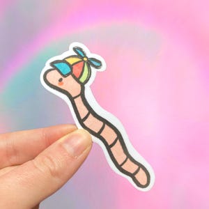 May include: A pink worm wearing a colorful hat with a propeller. The worm is outlined in black and has a white background.